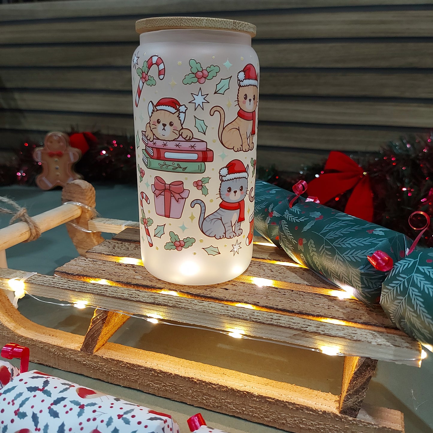 Festive frosted glass tumbler with cute Christmas cats, bamboo lid, and glass straw, on holiday display with lights and wrapping.